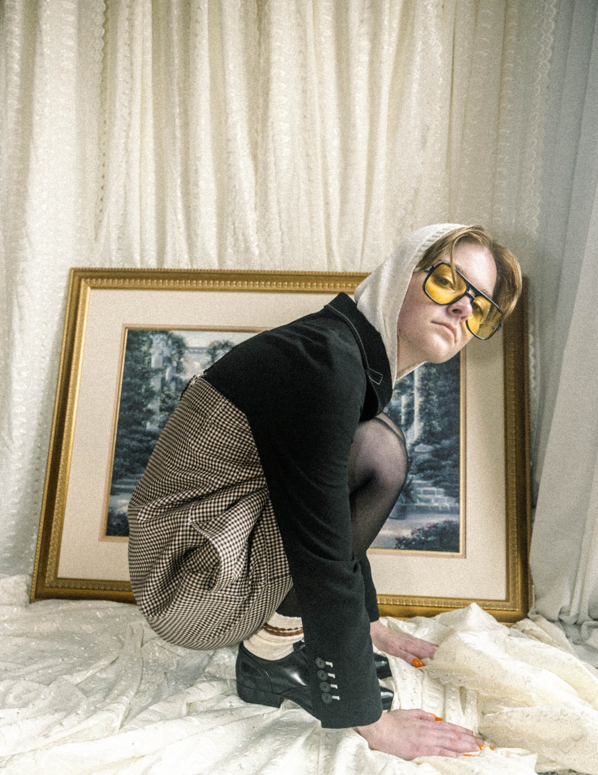 Queer fashion stylist Kati Kons squats side profile in a blazer and romper with hoodie and yellow sunglasses.