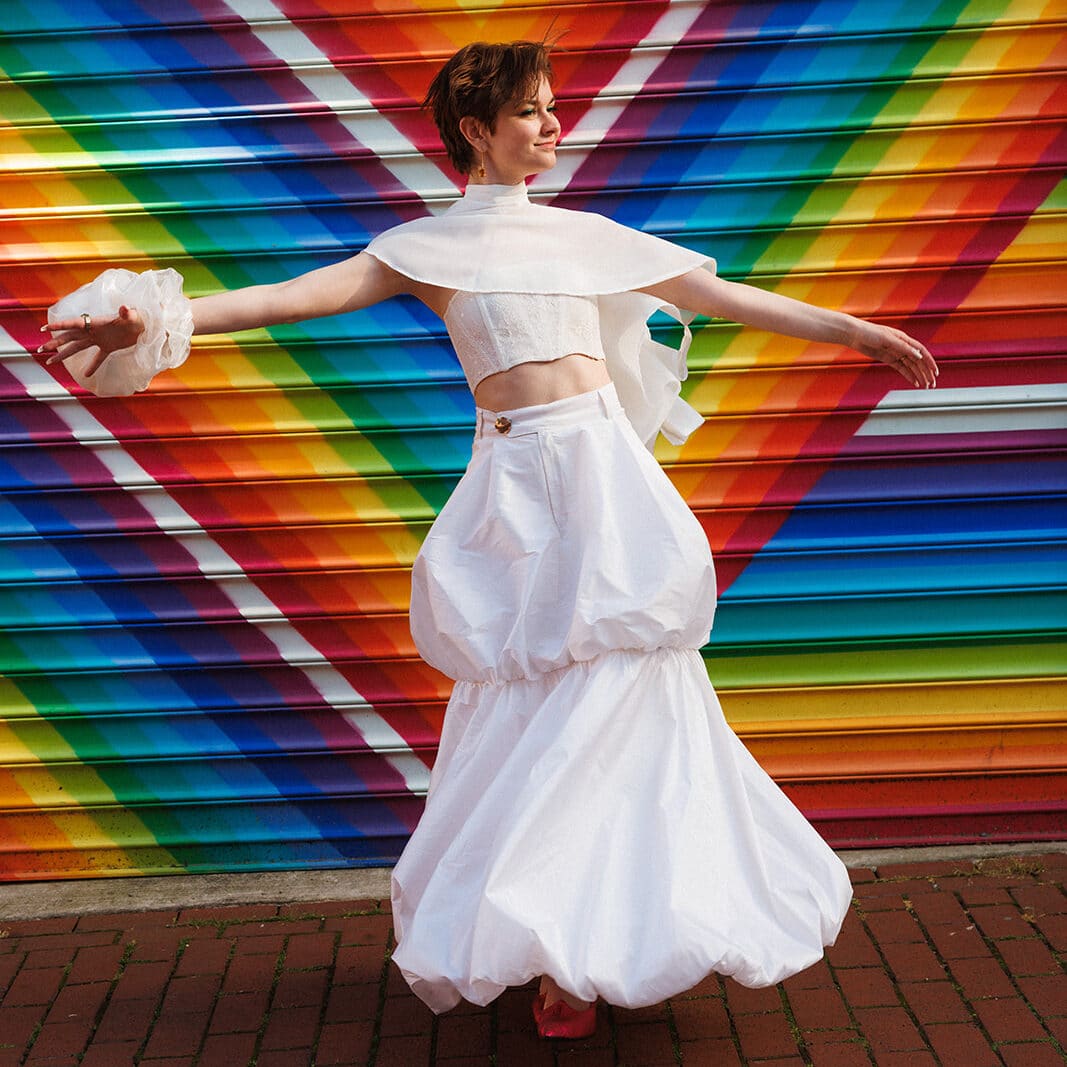 Kati Kons in a bustier top and capelet and double bubble hem skirt, all white, spinning around in front of a rainbow painted garage door.