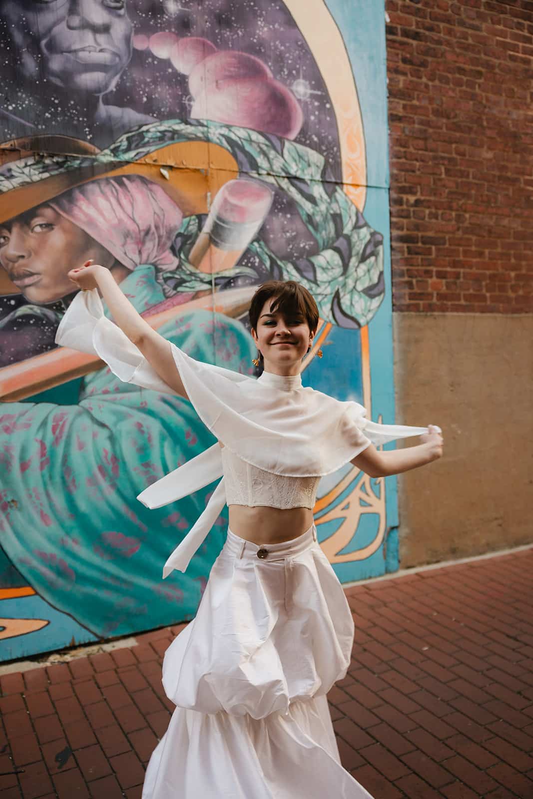 Picture of Queer fashion stylist Kati Kons spinning in a white bubble skirt and white bustier top and white capelet with a graffiti wall behind them.