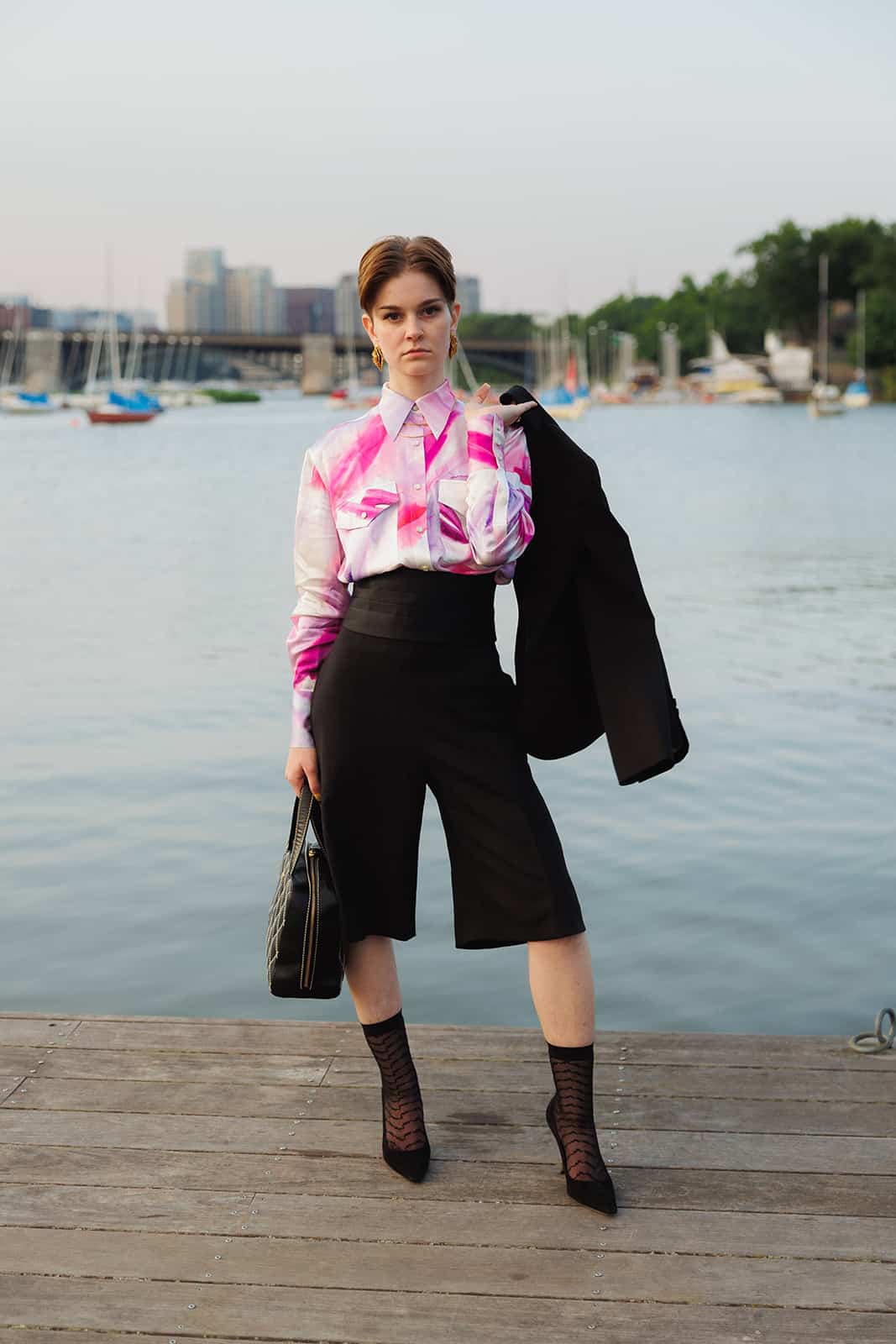 Queer Personal Stylist Kati Kons stands on a dock in Boston wearing a black shorts suit set with the jacket hanging over her shoulder and a bowler bag in the other hand. Styled with a pink and purple silk painterly shirt and a black silk cummerbund.
